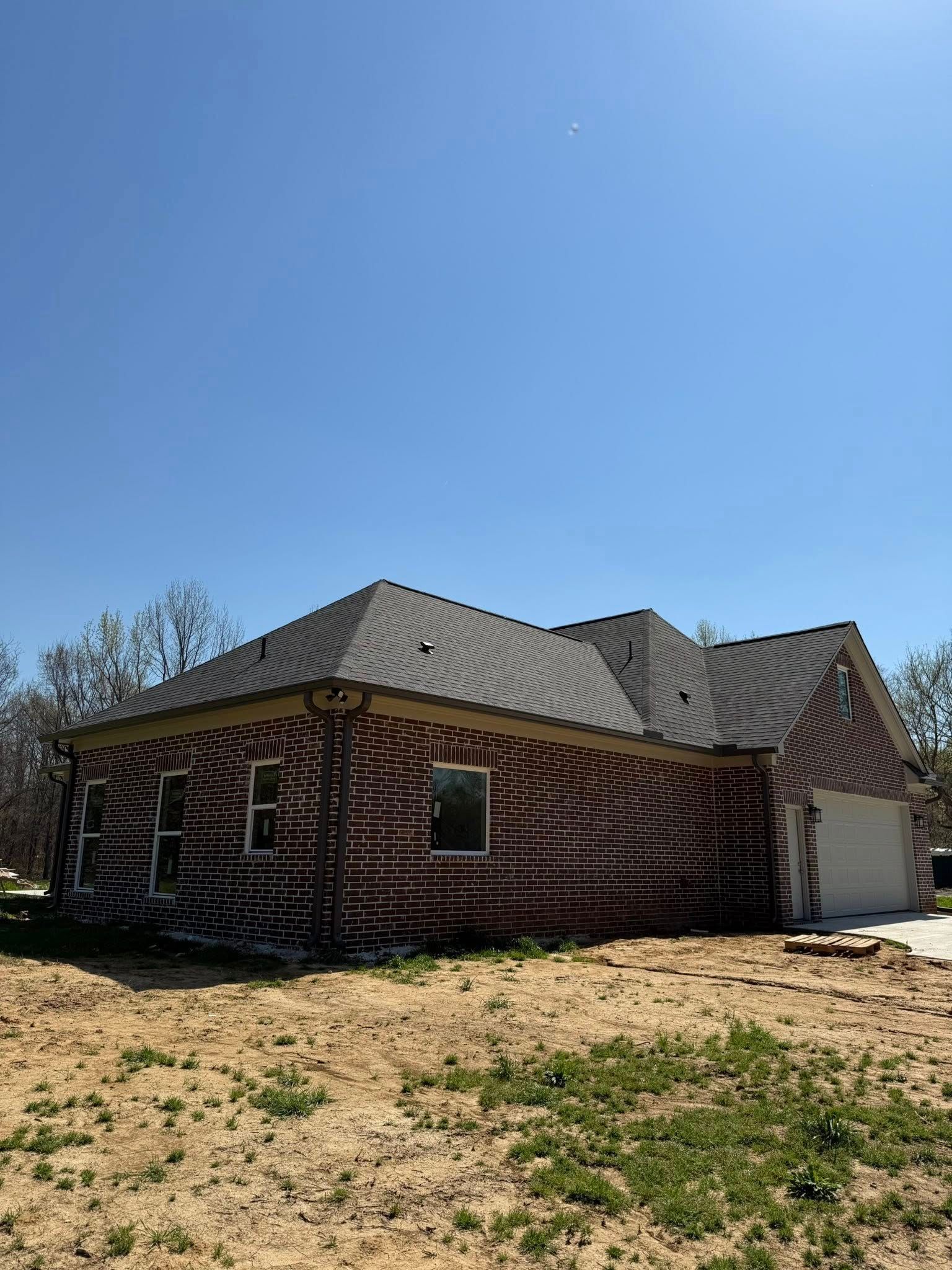 Brick house under construction with gray roof and bare dirt yard on a clear day