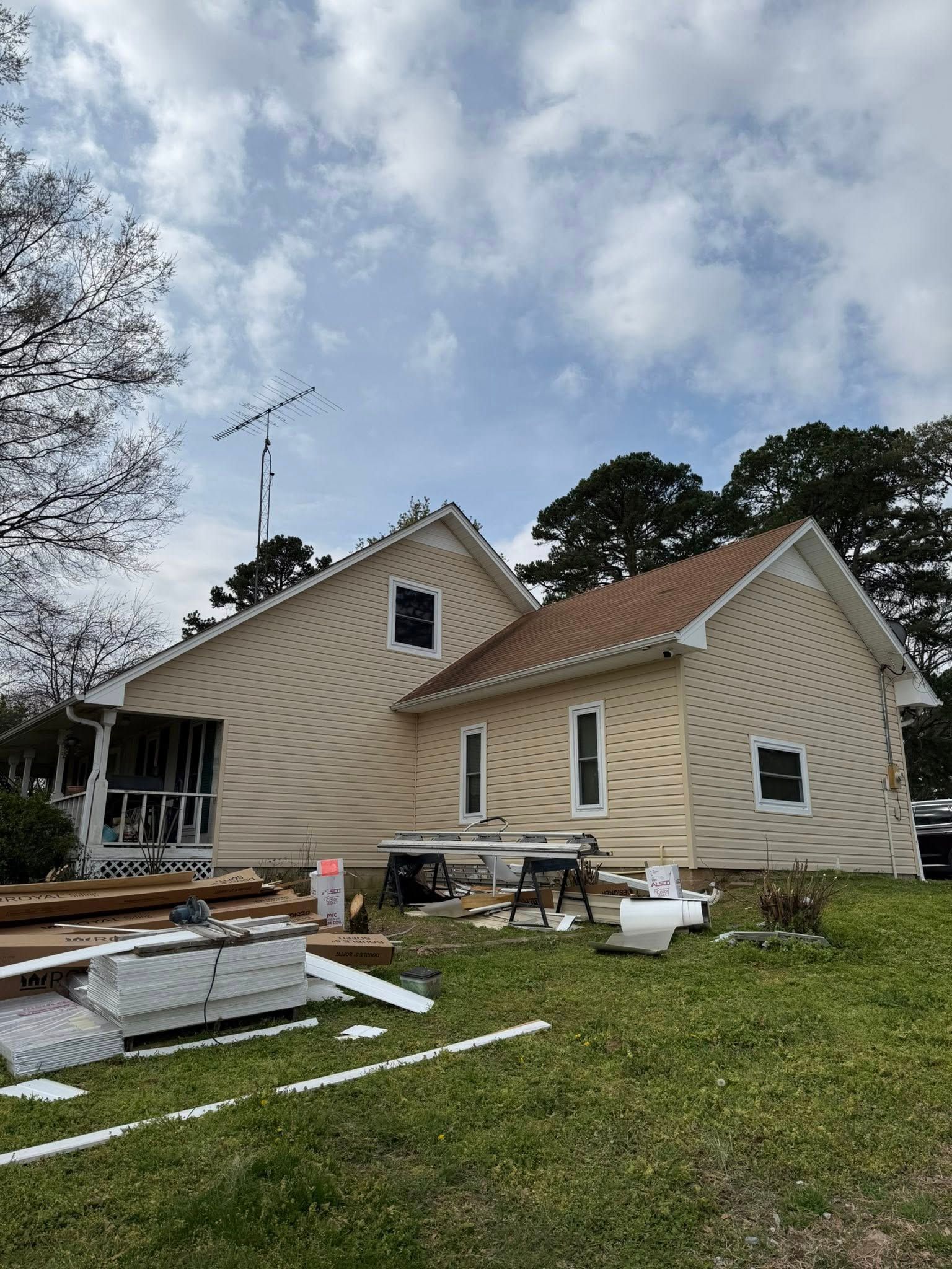 Damaged beige house with roof debris and construction materials scattered across the front lawn under a cloudy sky