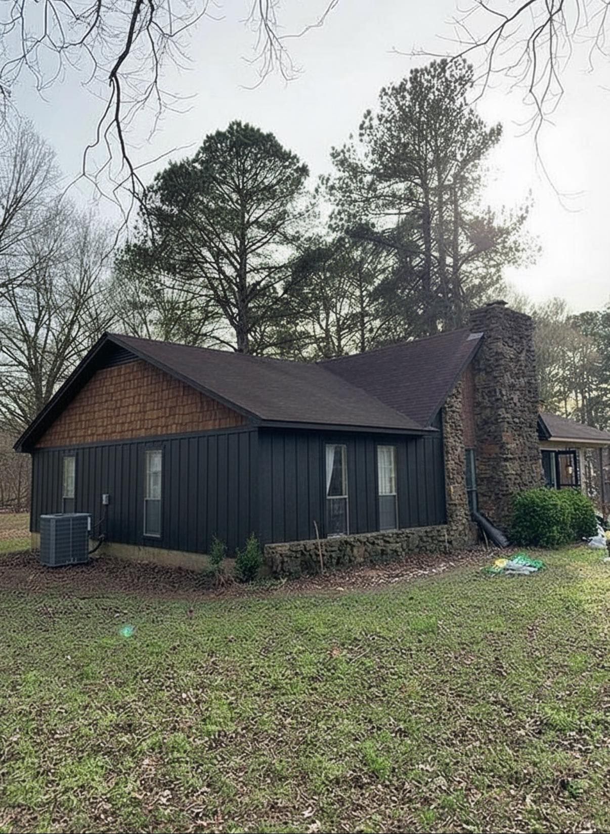 Small dark house with a brick chimney in a grassy yard under tall trees on an overcast day