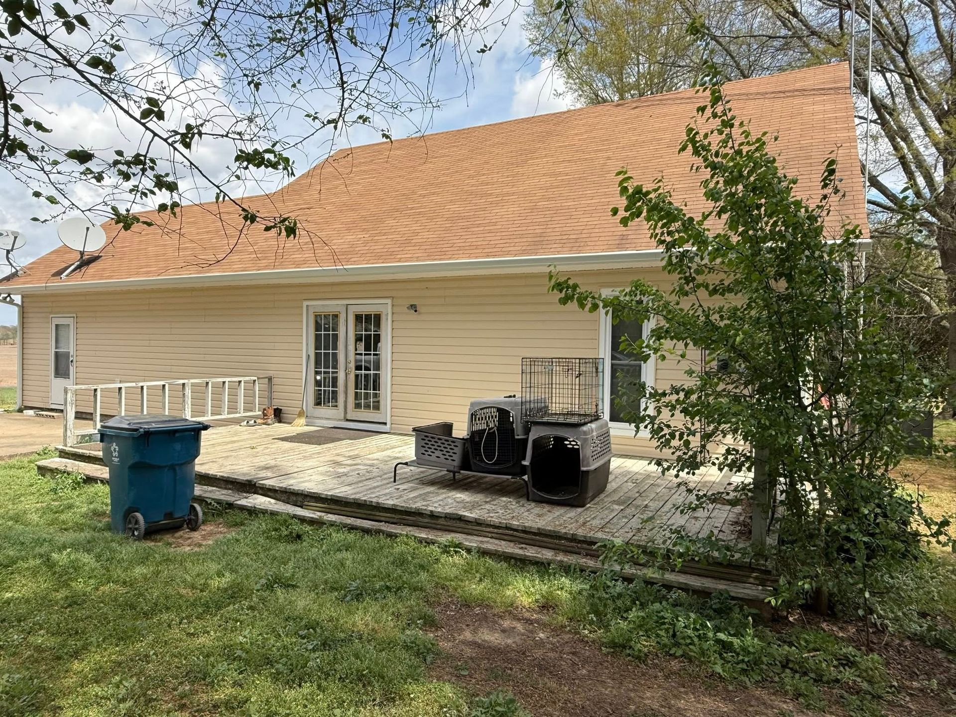 Single-story tan house with a large orange roof, concrete patio, grill, and blue trash bin in a backyard.