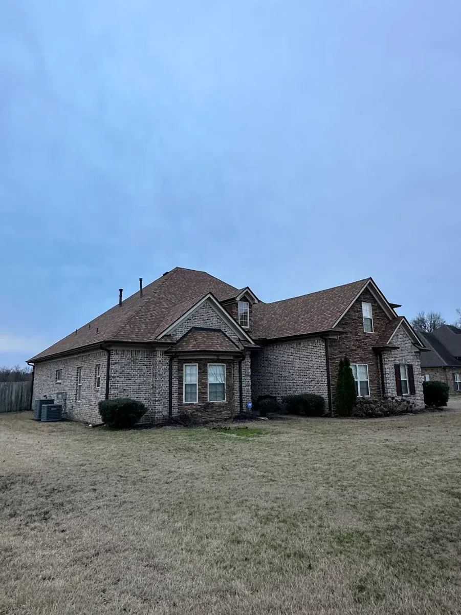 Stone house with a brown shingled roof on a grassy lot under an overcast sky.