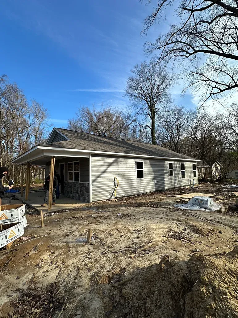 Single-story white house under construction on a muddy lot with leafless trees and blue sky