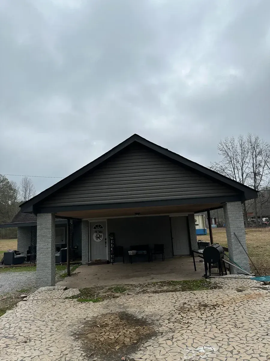 Single-story house with a covered carport, stone columns, and a gravel driveway under a cloudy sky