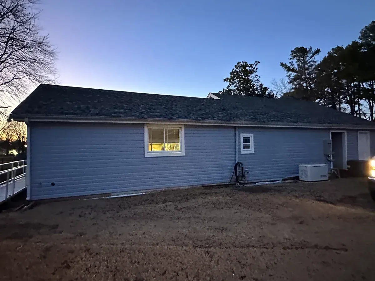 Single-story gray house at dusk with one lit window and a dirt yard.