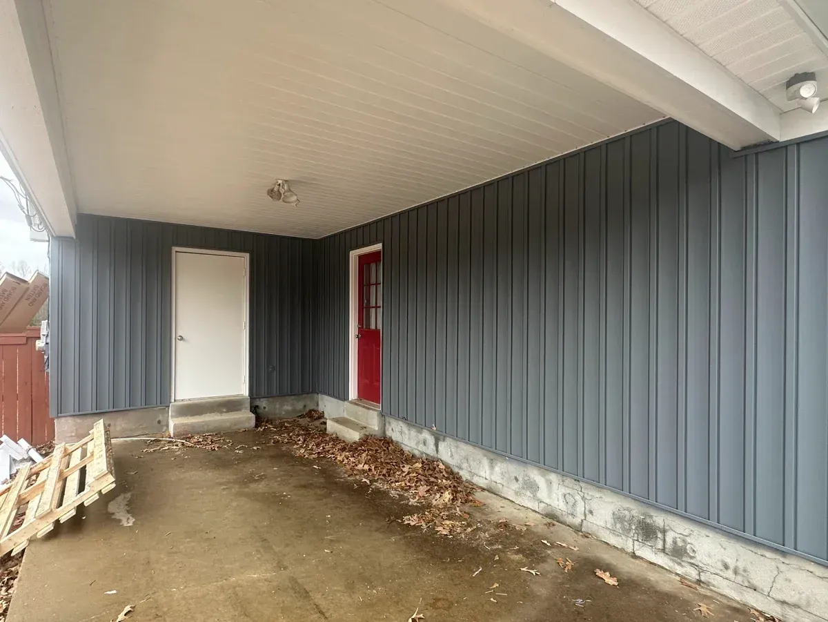 Covered side porch with gray siding, white ceiling, red door, and dirt ground.