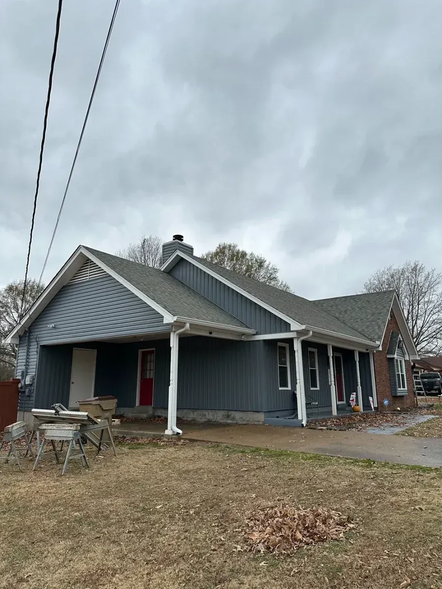 Gray house with a gabled roof and front porch under a cloudy sky, with bare yard and chairs outside