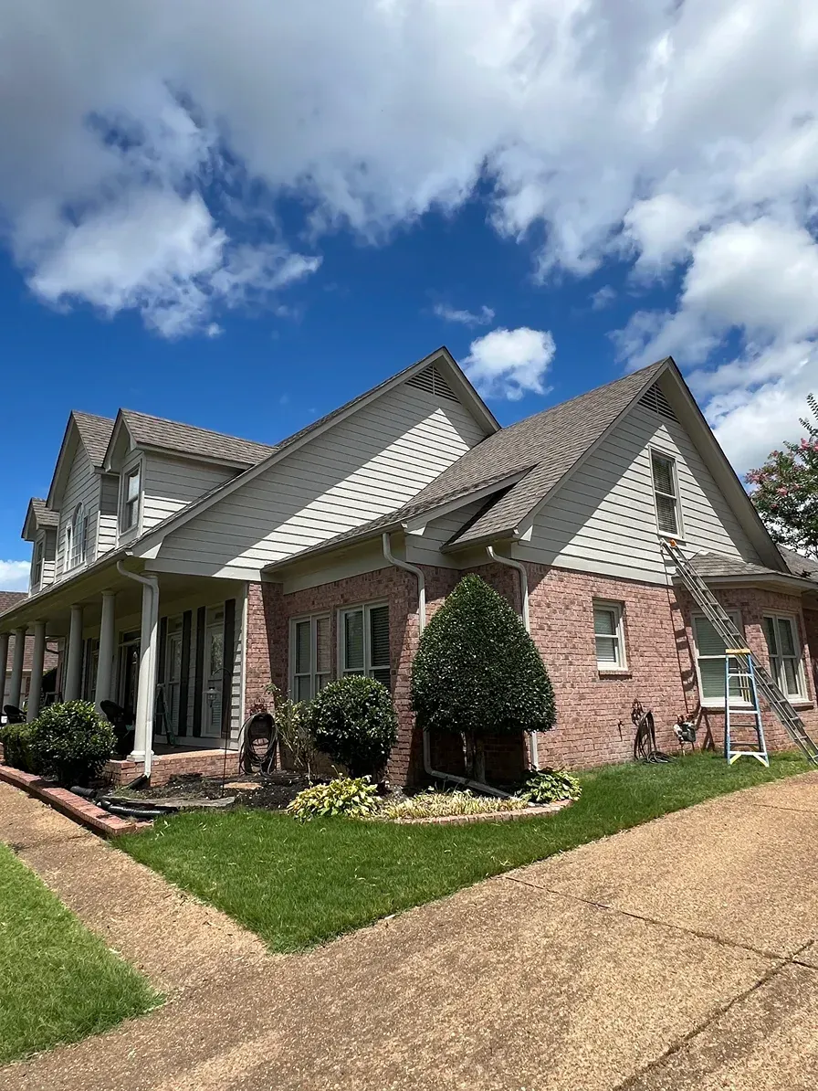 Brick suburban house with gray roof, white trim, and a curved driveway under a blue, partly cloudy sky