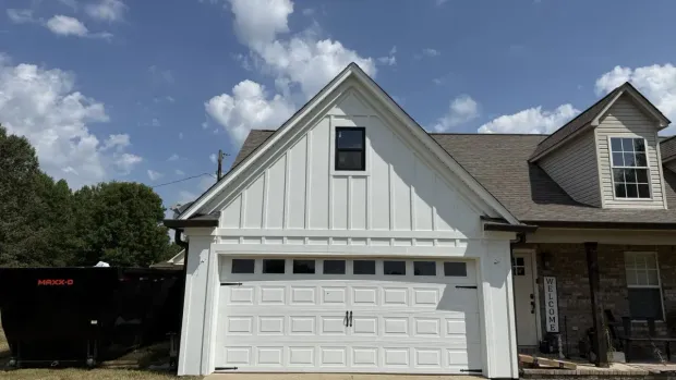 White gabled house front with two-car garage and dormer windows under a blue sky