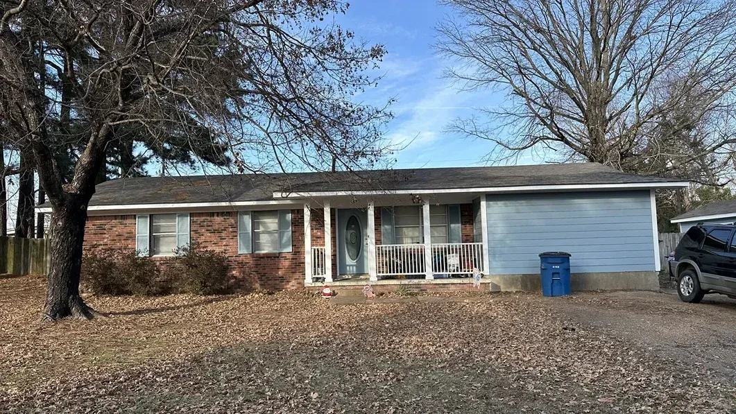 Single-story brick house with a blue trash bin, leaf-covered yard, and a parked vehicle on the right.