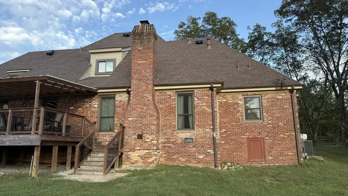 Rear view of a brick-and-wood house with a chimney, deck, and grassy yard under a cloudy sky