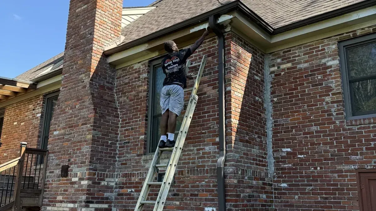 Person on a ladder cleaning a brick house exterior near a window and chimney