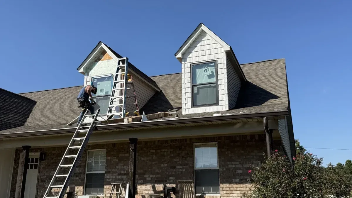 Person on a ladder painting a dormer window on a brick house under a blue sky