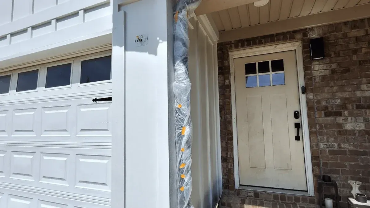 Garage and front door of a house with white siding, brick exterior, and a damaged door frame wrapped in plastic.