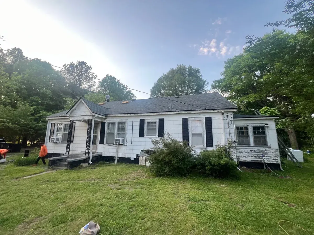 White farmhouse with peeling paint and black shutters on a grassy yard, surrounded by trees under a bright sky