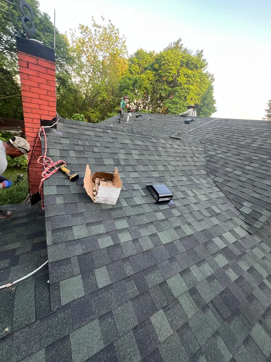 Rooftop with dark gray shingles, red brick chimney, and a white vent box under trees at sunset