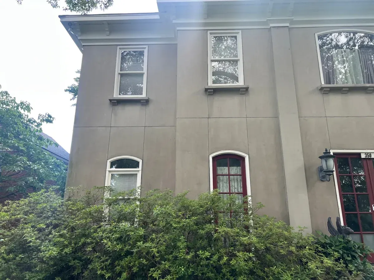 Gray two-story house with barred windows and overgrown bushes in front