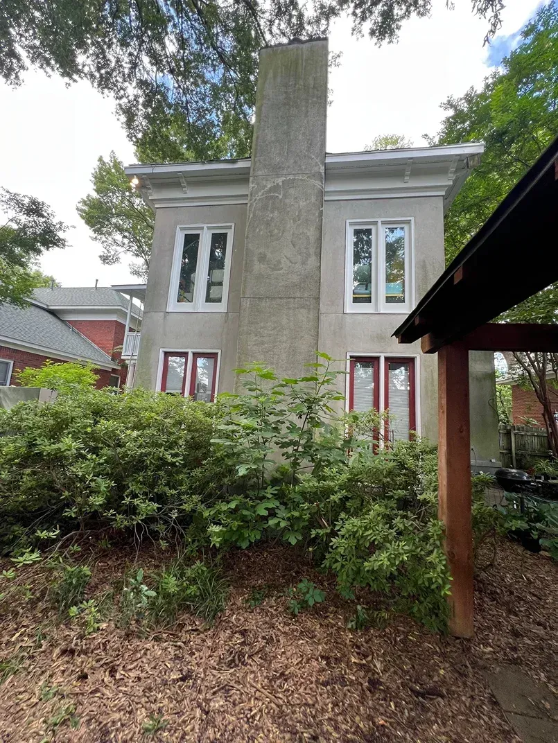 Two-story white house with a tall central chimney, red doors, and overgrown shrubs in front