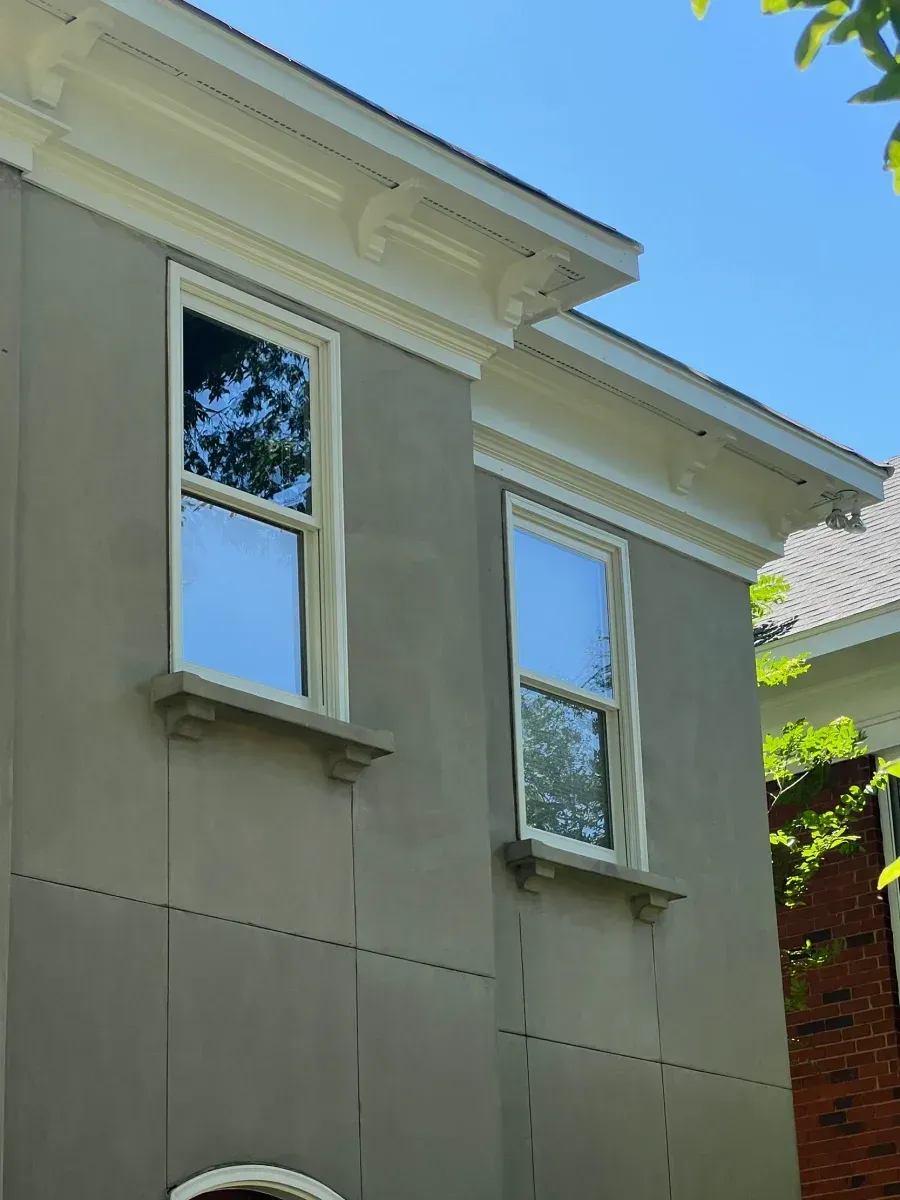 Beige house exterior with two white-trimmed windows and decorative roof molding under a blue sky
