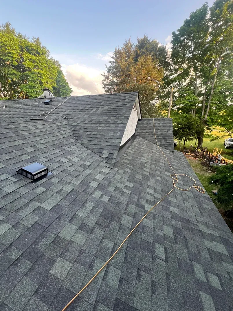Roof covered in dark gray shingles with a vent, beside trees and a blue sky