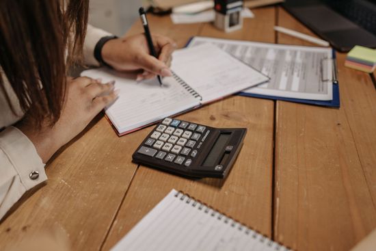 Hands writing on a notepad with a calculator and papers on a wooden table