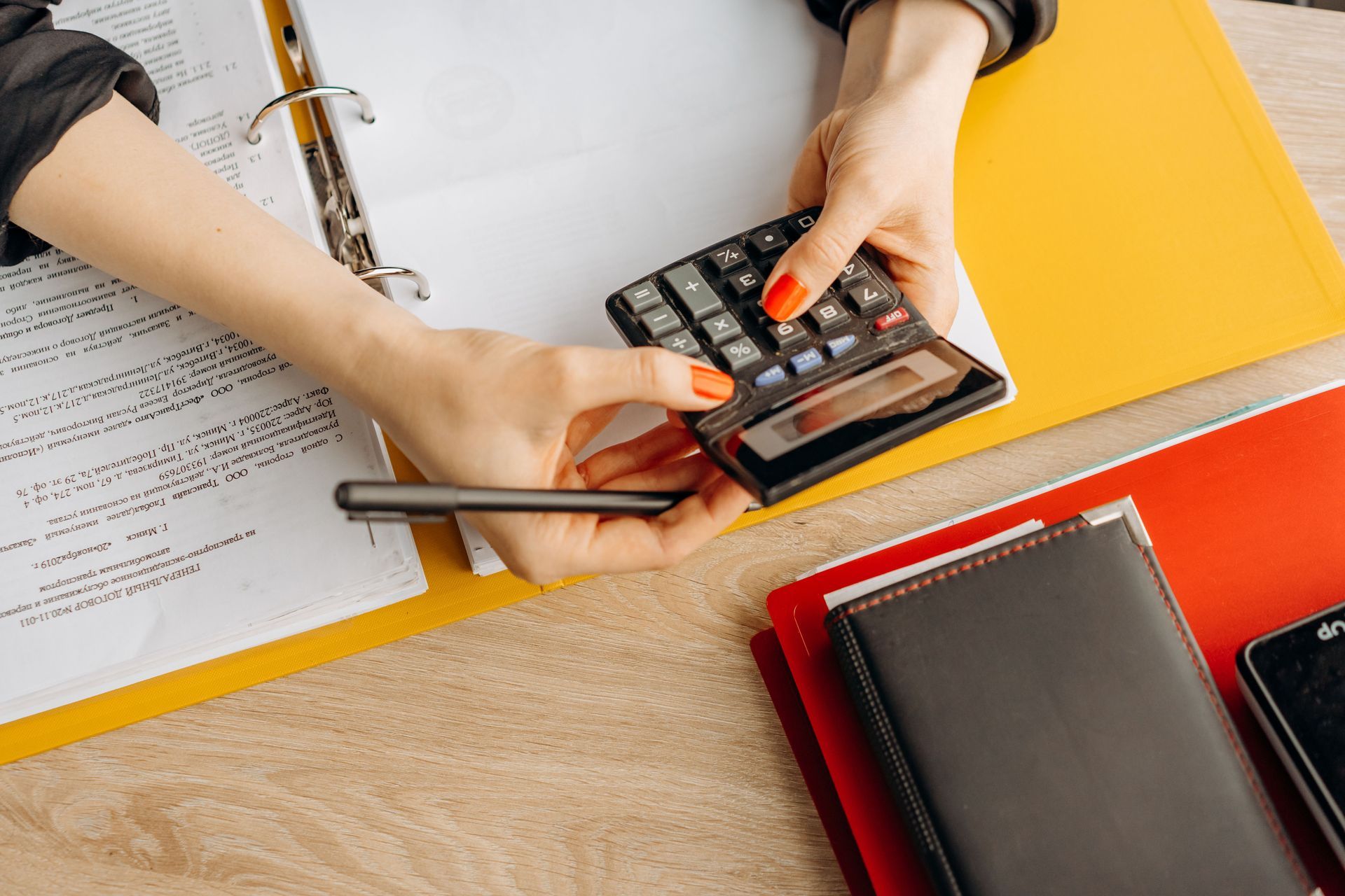 Hands using a smartphone and calculator over papers on a desk with orange and red folders