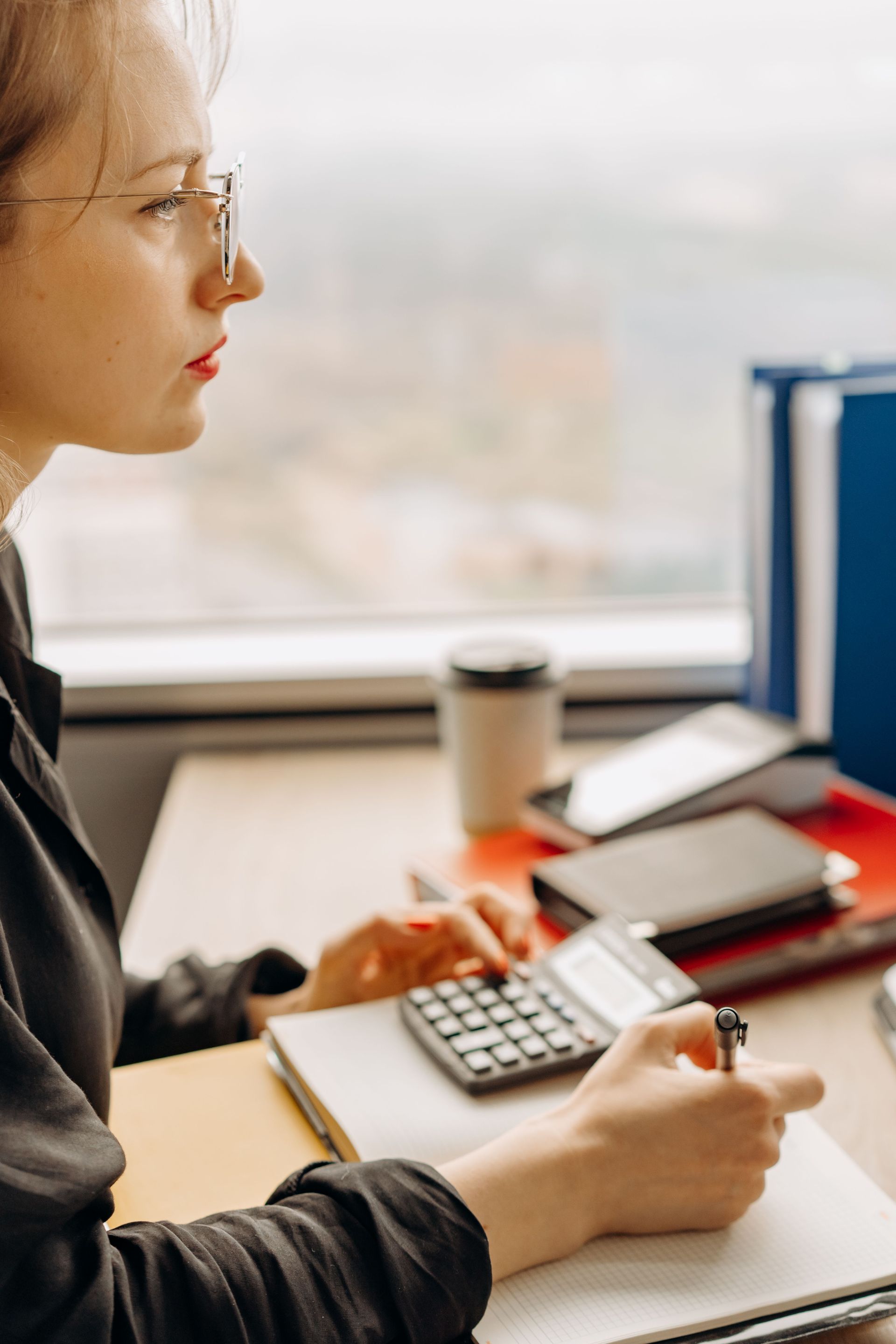 Person writing in a notebook at a desk with a calculator, coffee cup, and laptop nearby