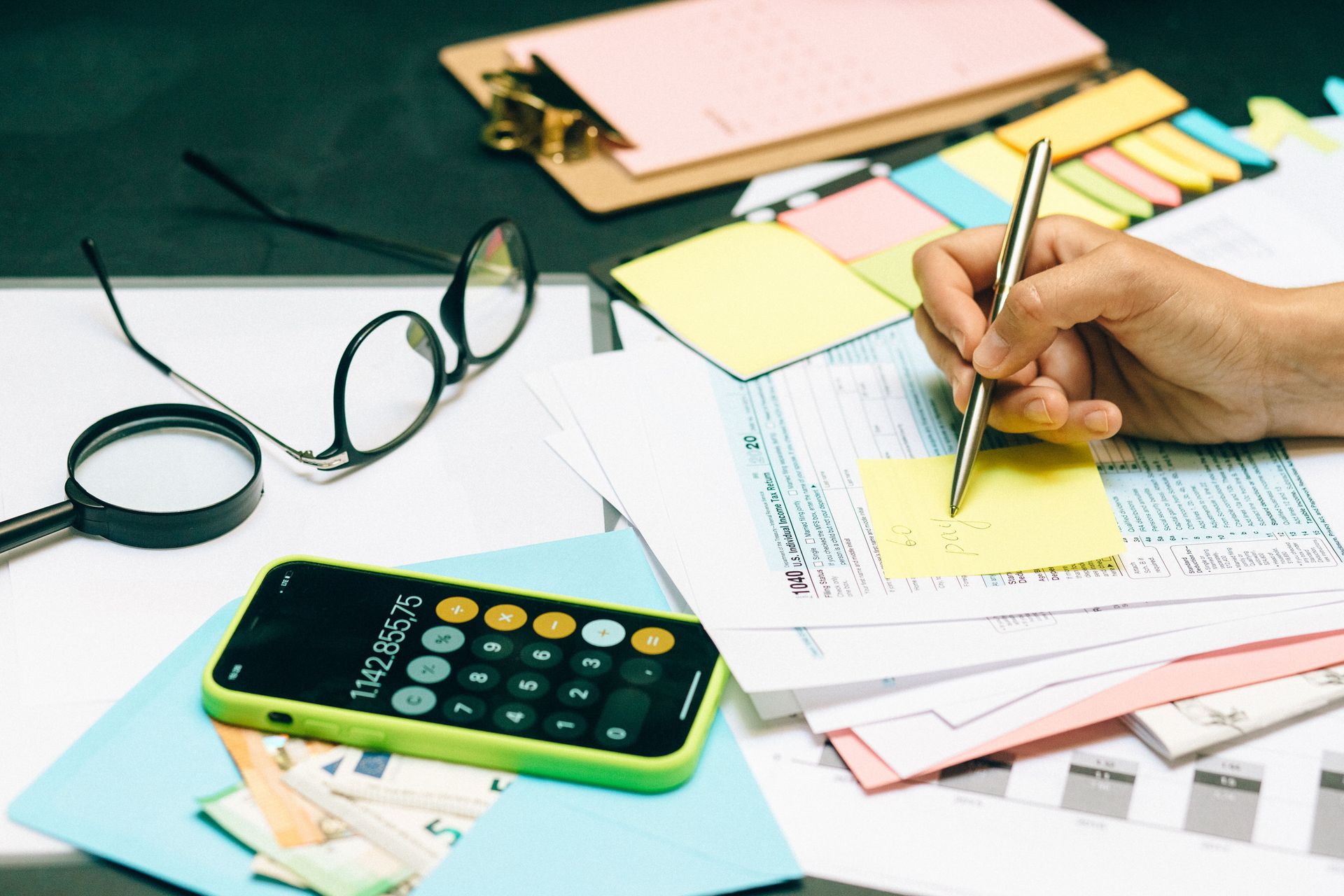 Hand writing on a financial form beside a calculator, glasses, and sticky notes on a desk