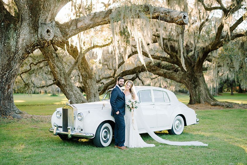 A bride and groom are standing next to a white car in a field.