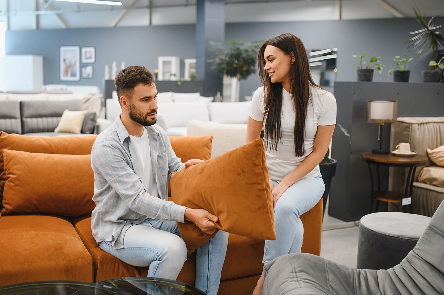 Two people sitting on a sofa in a furniture store, examining an orange throw pillow together. Two people sitting on a sofa in a furniture store, examining an orange throw pillow together.