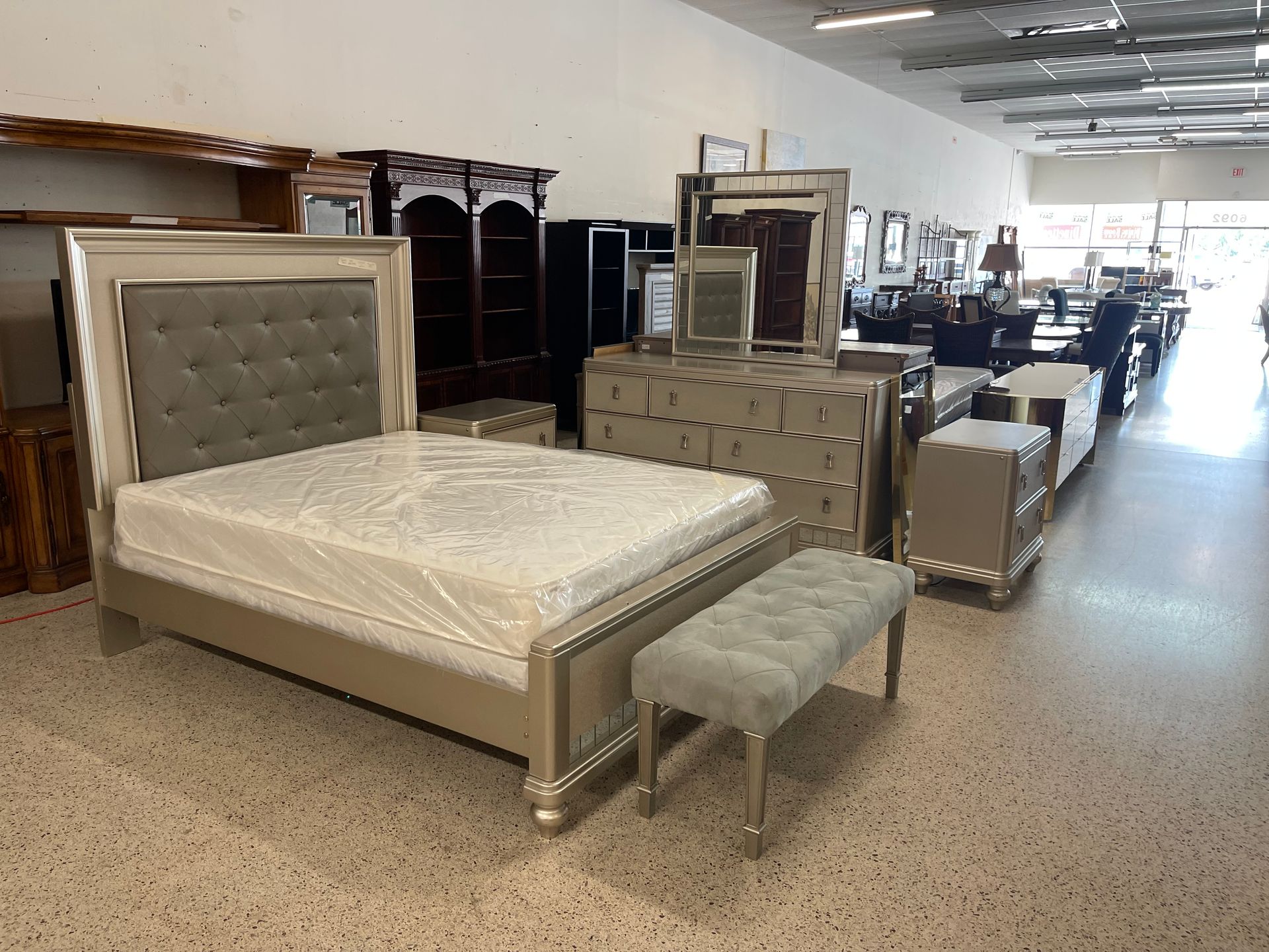 A furniture showroom floor displays a tufted gray bedroom set, including a bed, matching bench, dresser, and nightstand.