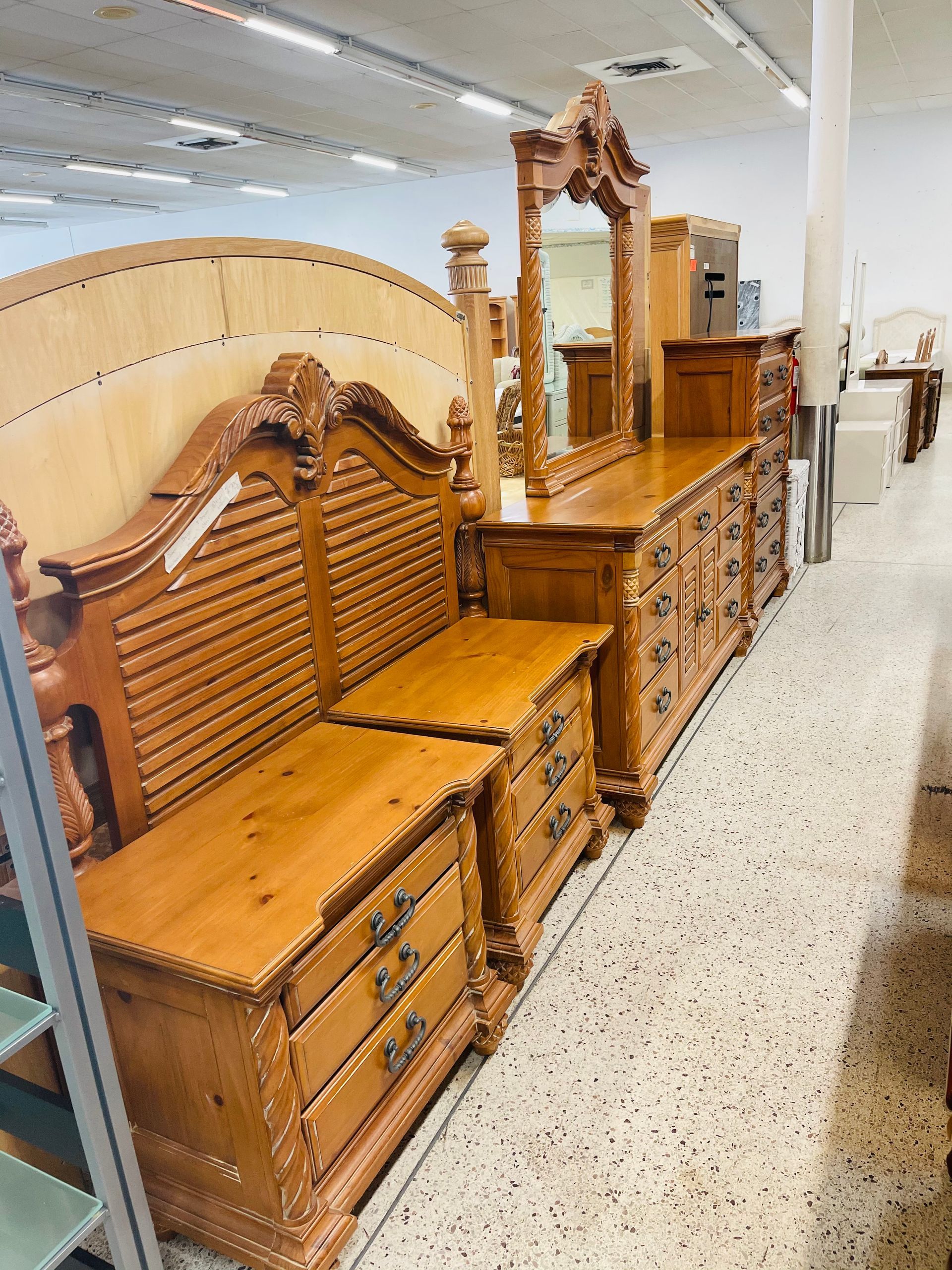 Ornate, light-brown wood bedroom furniture including a headboard, nightstands, and a dresser with mirror in a store.