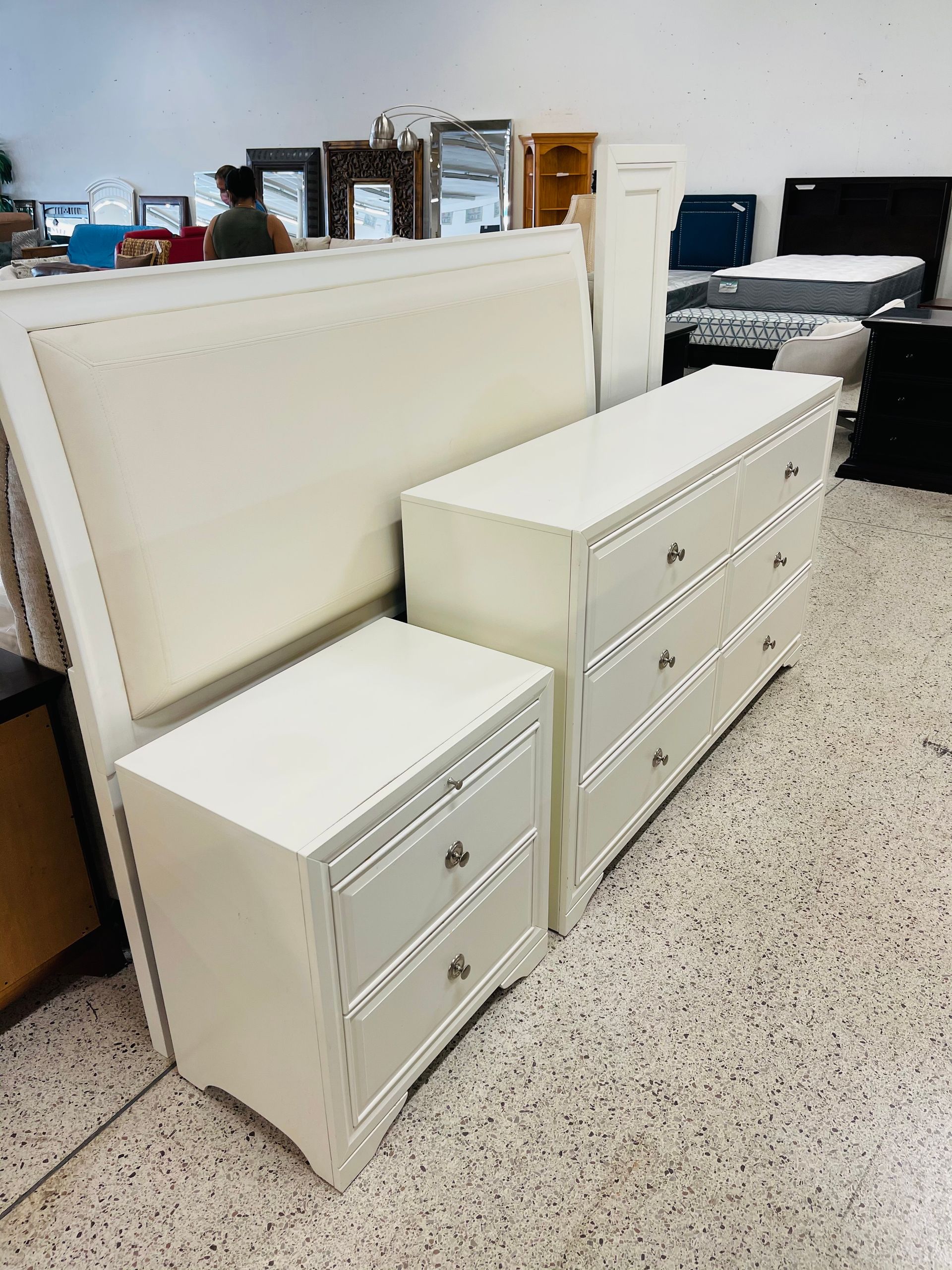 A white bedroom furniture set featuring a large headboard, a two-drawer nightstand, and a long dresser in a showroom.