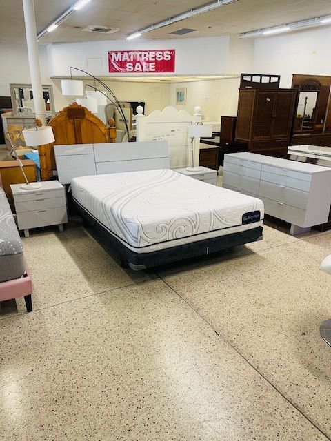 A mattress display in a furniture store featuring a white bed set, dresser, and lamps under a 