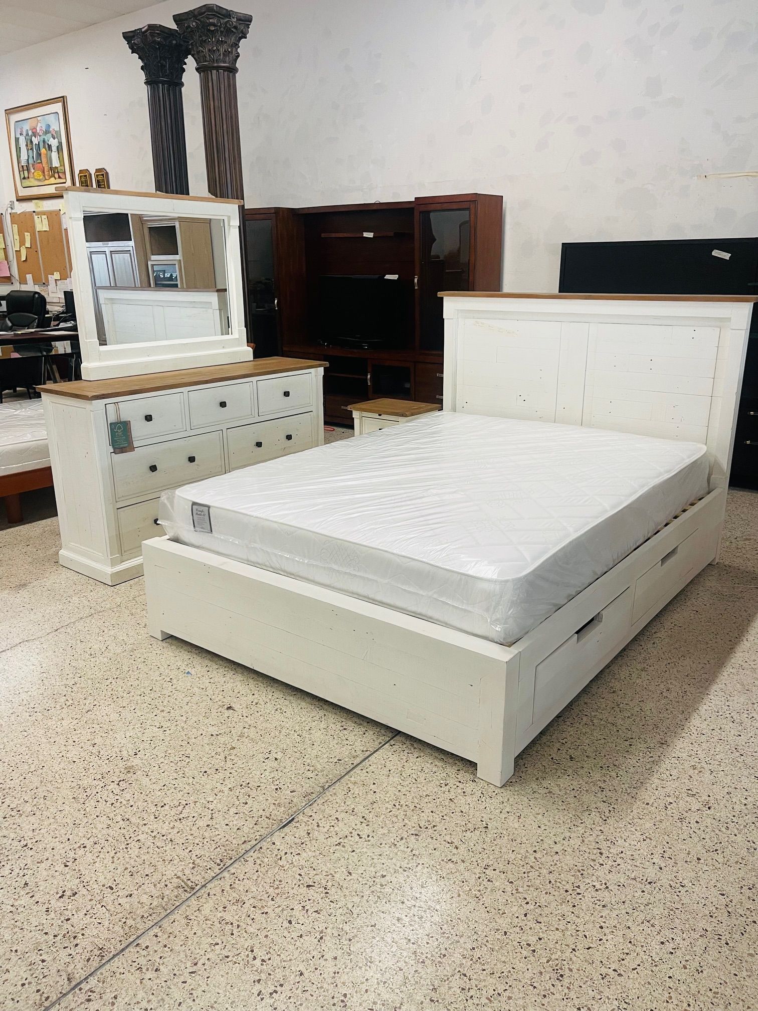 White bedroom furniture set, including a bed frame with storage drawers and a dresser with a mirror, in a showroom.