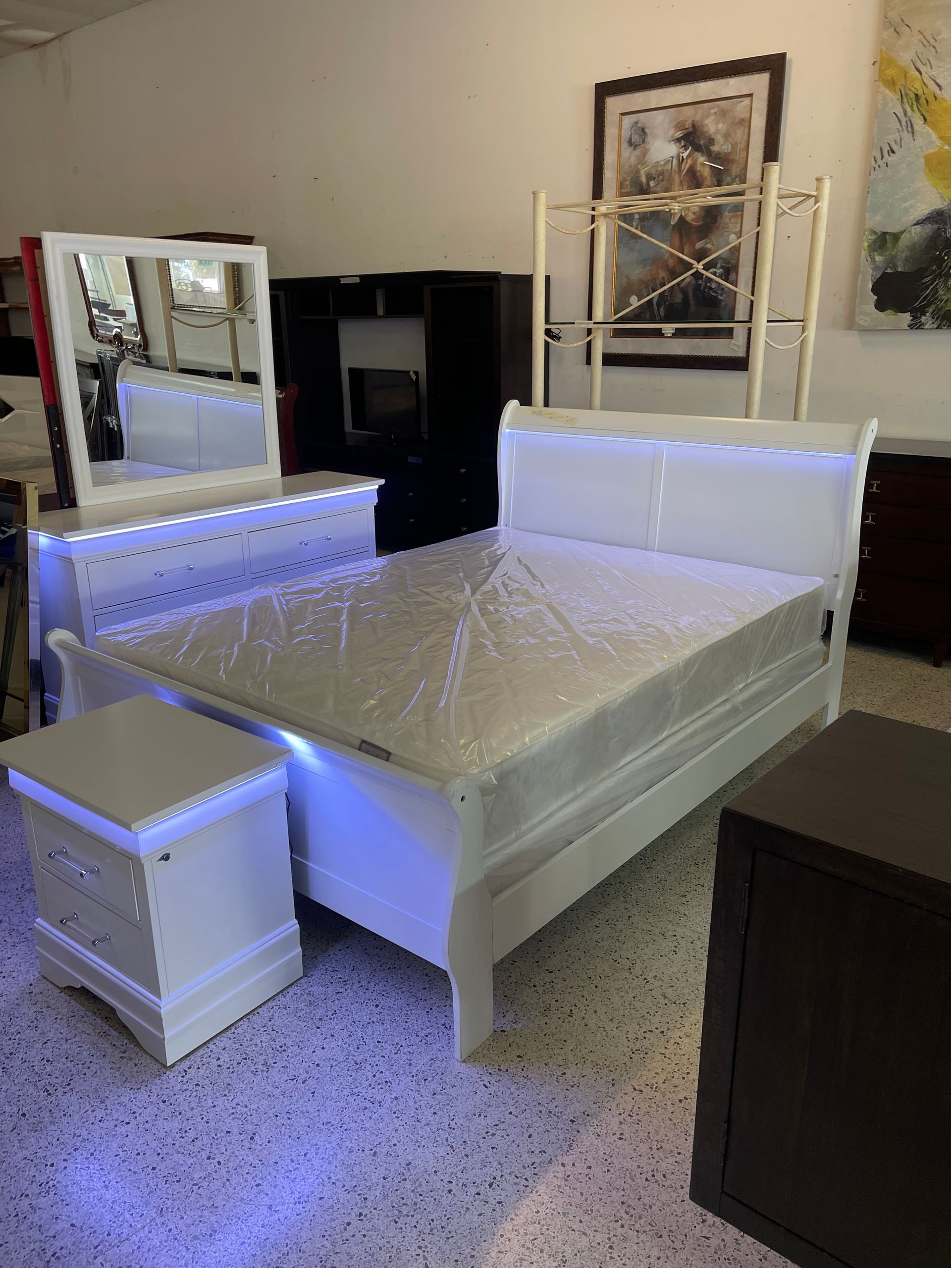 White bedroom set with an LED-lit headboard, a matching nightstand, and a dresser with a mirror, displayed in a showroom.