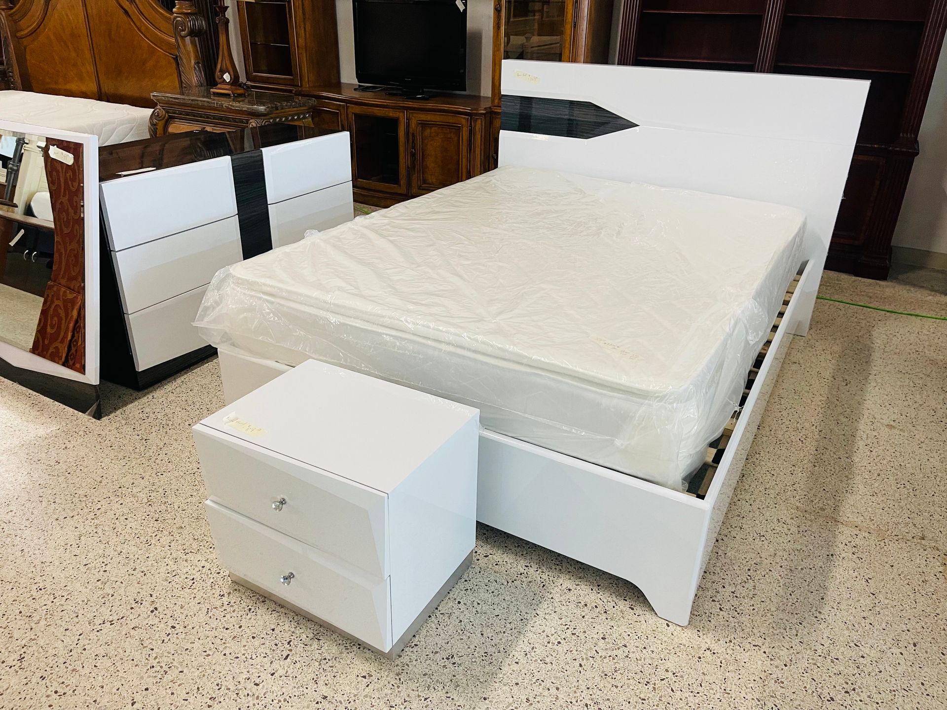 A white, modern double bed with a matching nightstand in a furniture showroom with speckled flooring.