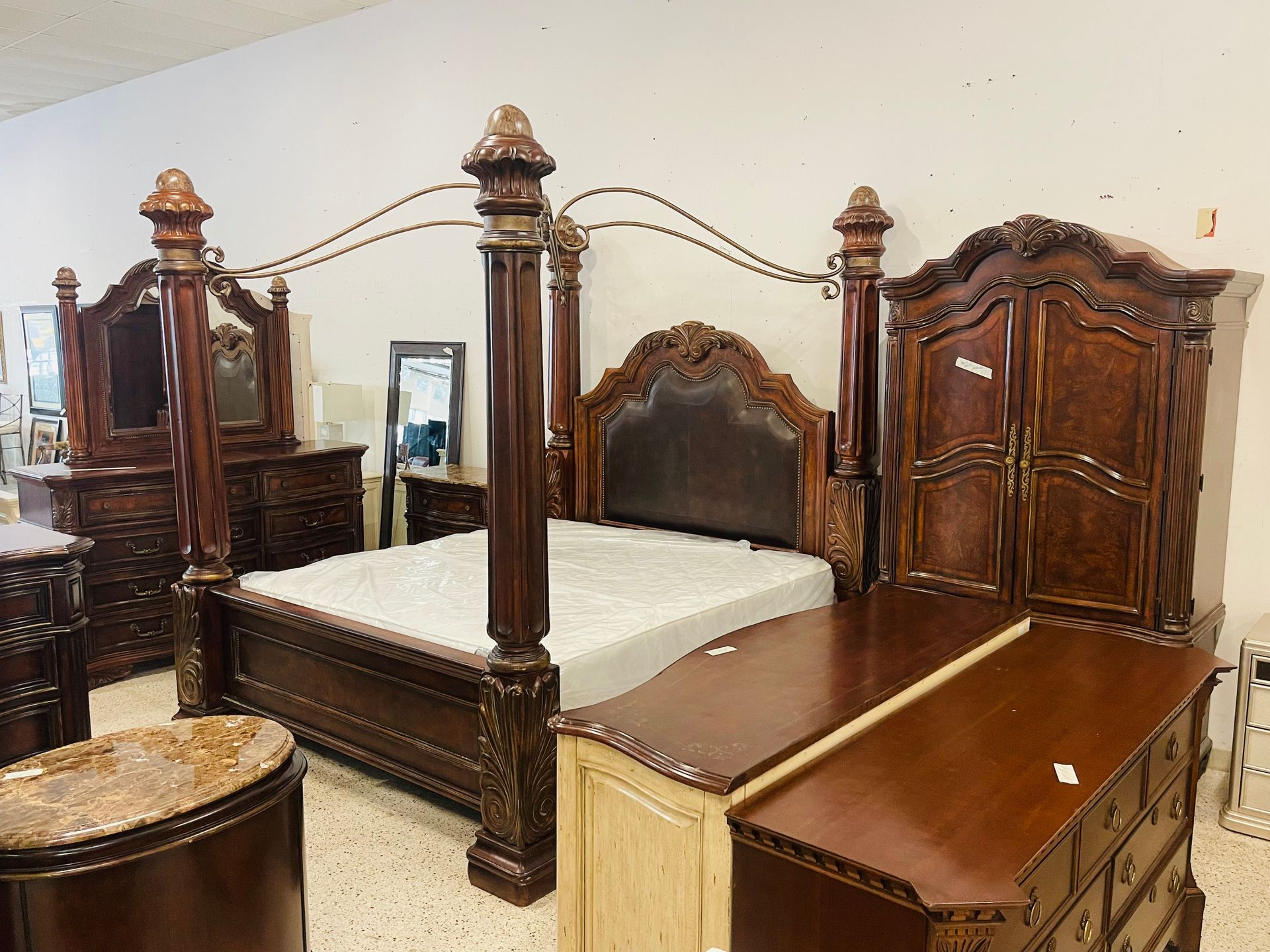 A showroom display featuring a large, ornate wooden four-poster canopy bed, a matching wardrobe, and bedroom dressers.
