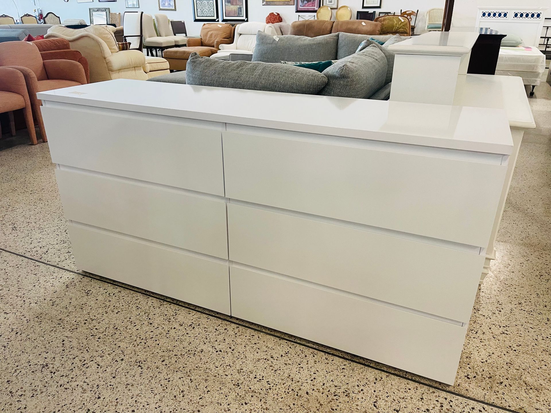 A white, six-drawer dresser with a glossy finish stands in a showroom with other furniture pieces in the background.