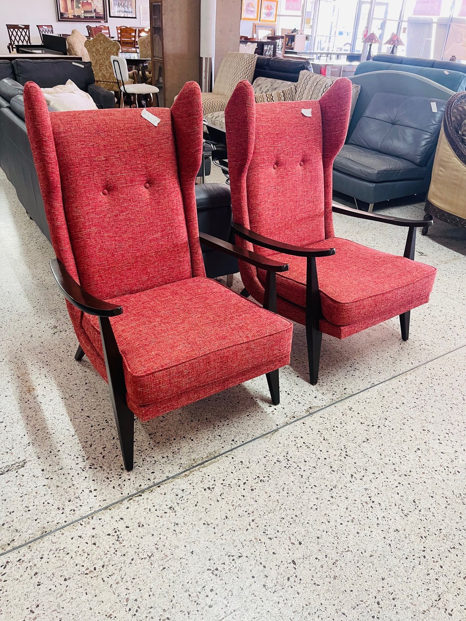 Two matching red wingback chairs with dark wooden arms and legs placed on a speckled tile floor in a furniture store.