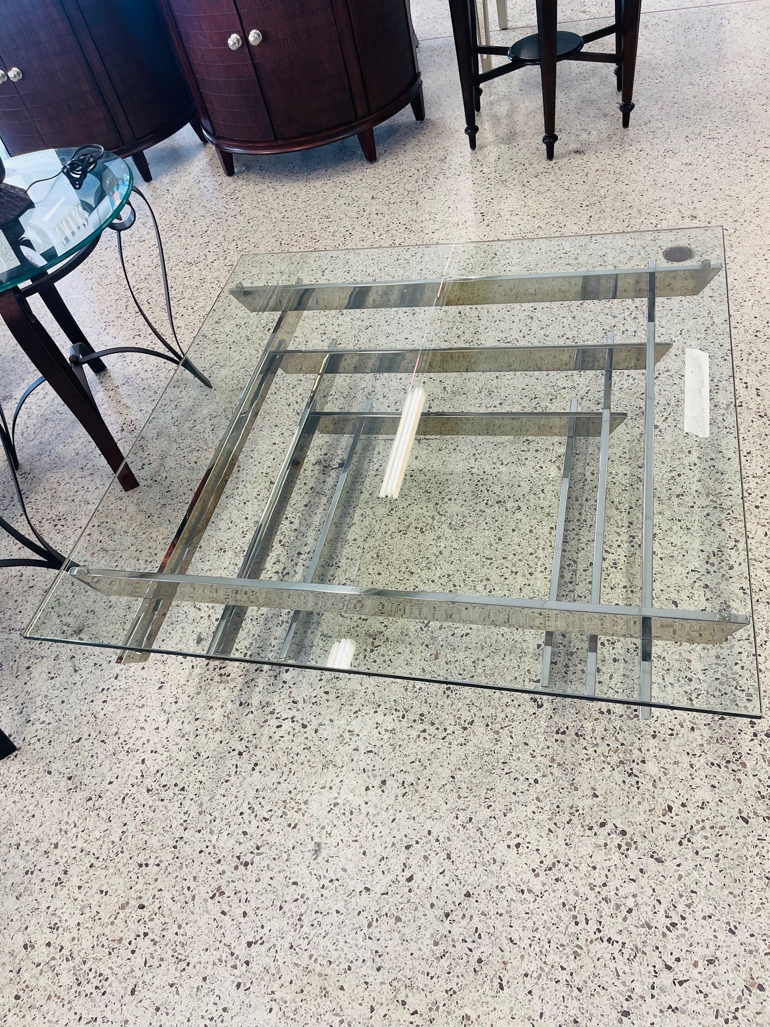 A clear glass coffee table with a geometric silver metal frame, positioned on a speckled floor in a store.