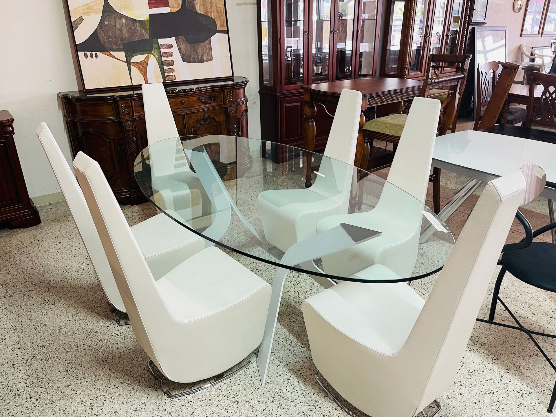 A glass-topped dining table with six modern, high-backed white chairs on a speckled floor in a furniture showroom.