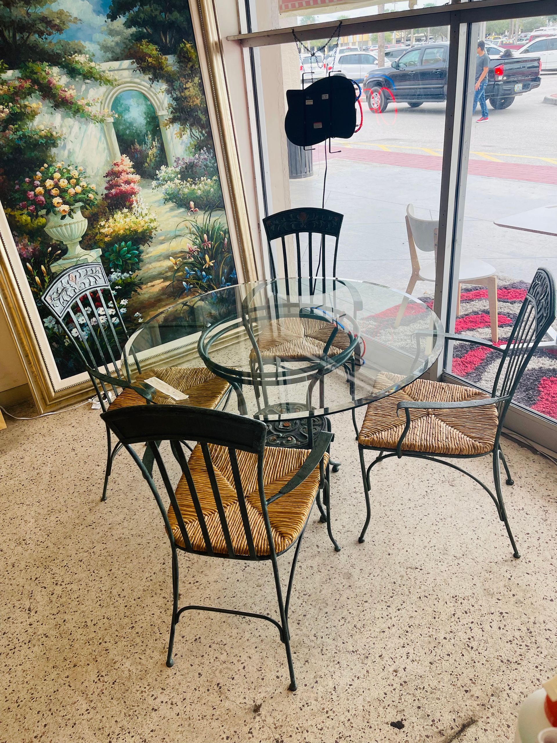A glass-topped table set with four black metal chairs with patterned cushions, arranged near a window in a shop.
