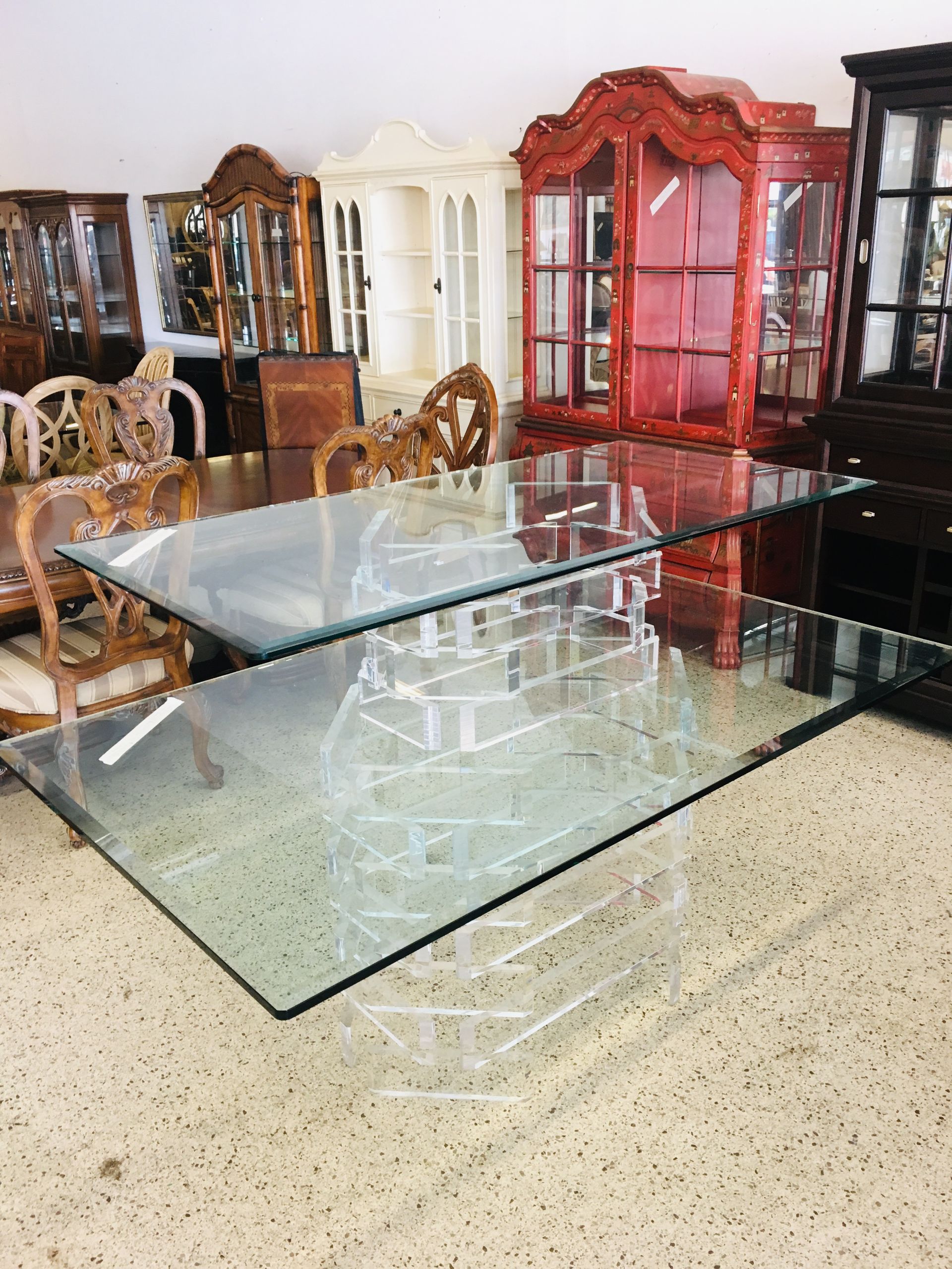 A glass dining table with a transparent acrylic base, displayed in a furniture showroom with various cabinets behind it.