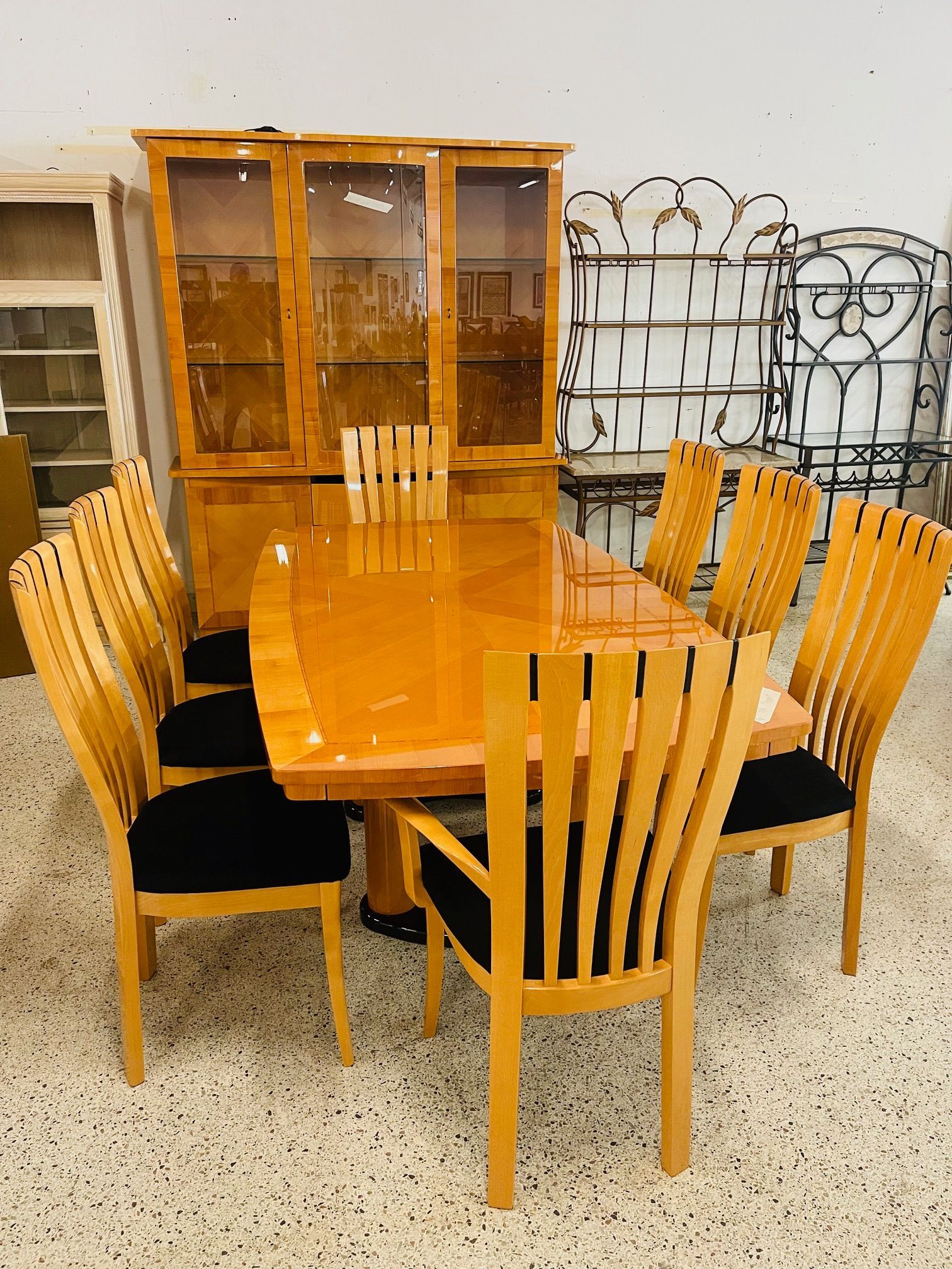 A glossy wooden dining set with a hutch, six chairs with black seats, and two metal decorative shelves in a bright room.