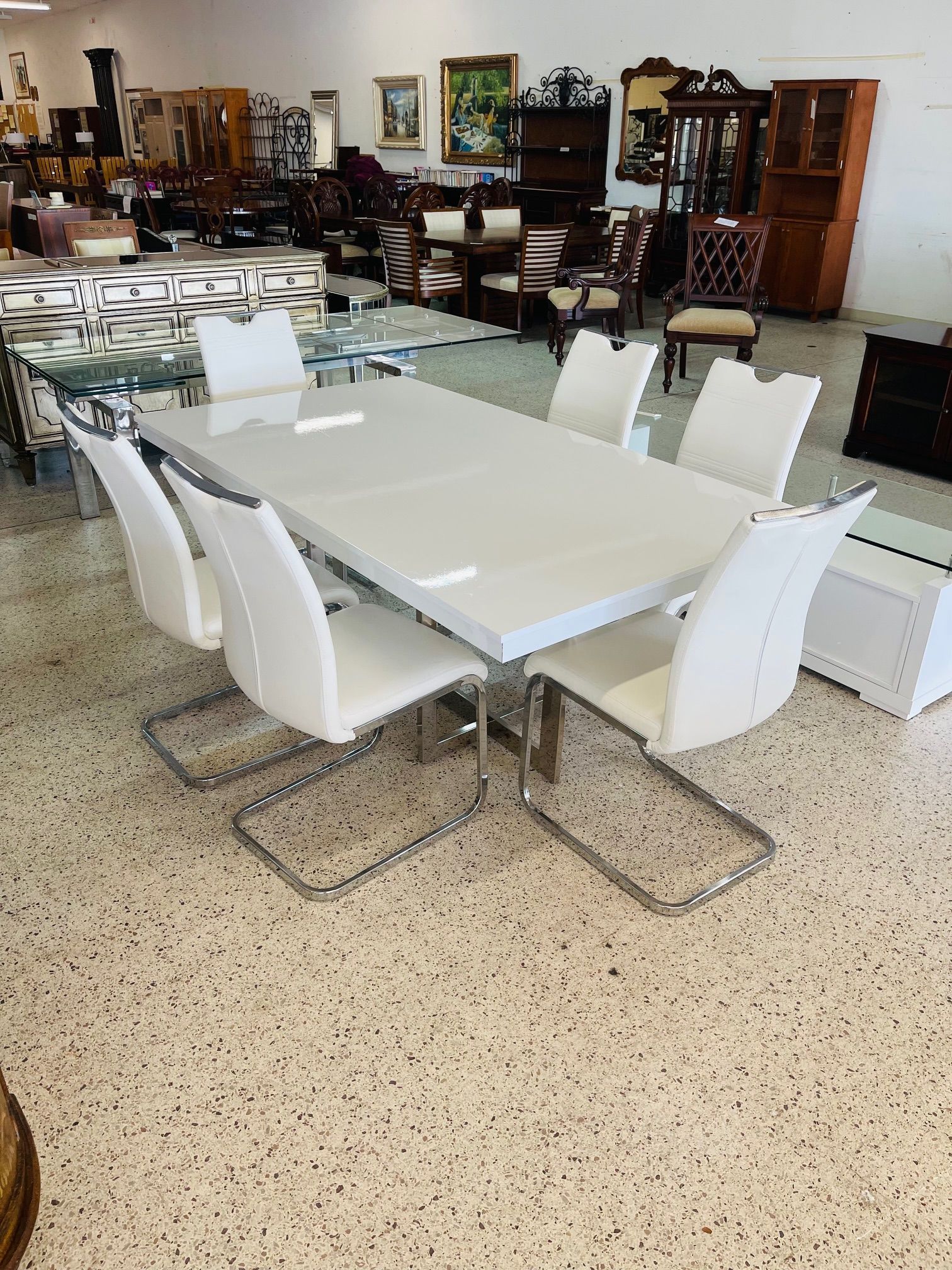 A modern white rectangular dining table with six white upholstered chairs on a speckled floor in a furniture store.