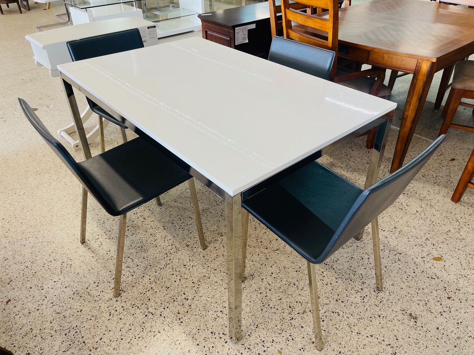 A rectangular white dining table with four modern, dark blue, high-backed chairs set on a speckled floor in a store.