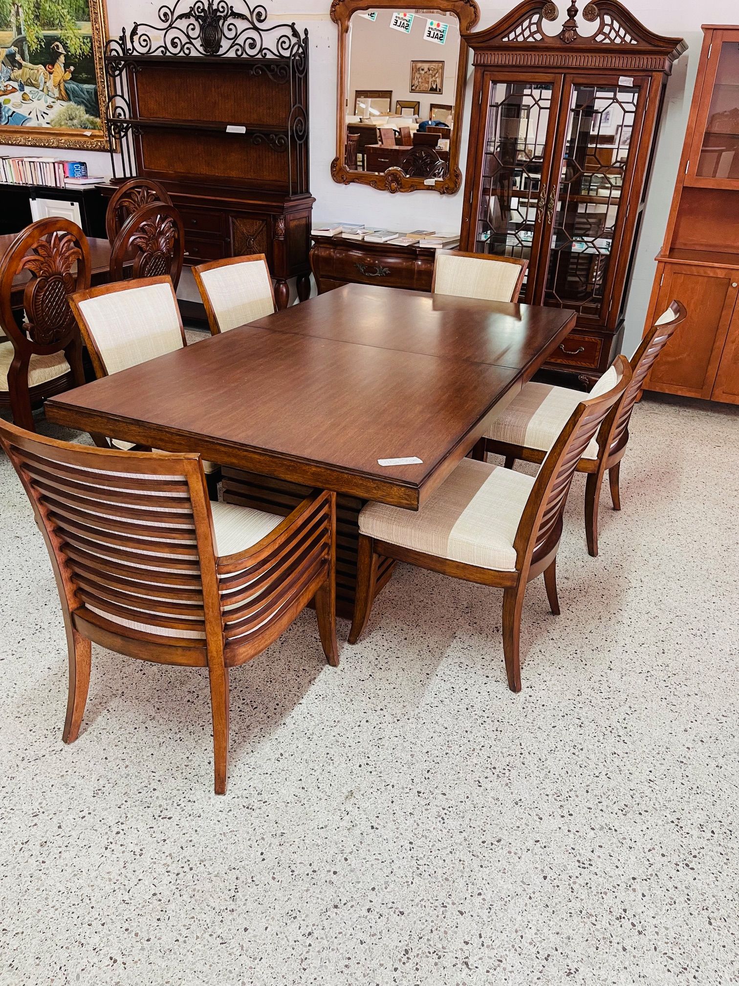 A wooden dining set with six chairs featuring slatted backs and beige cushions, arranged on a speckled floor in a store.