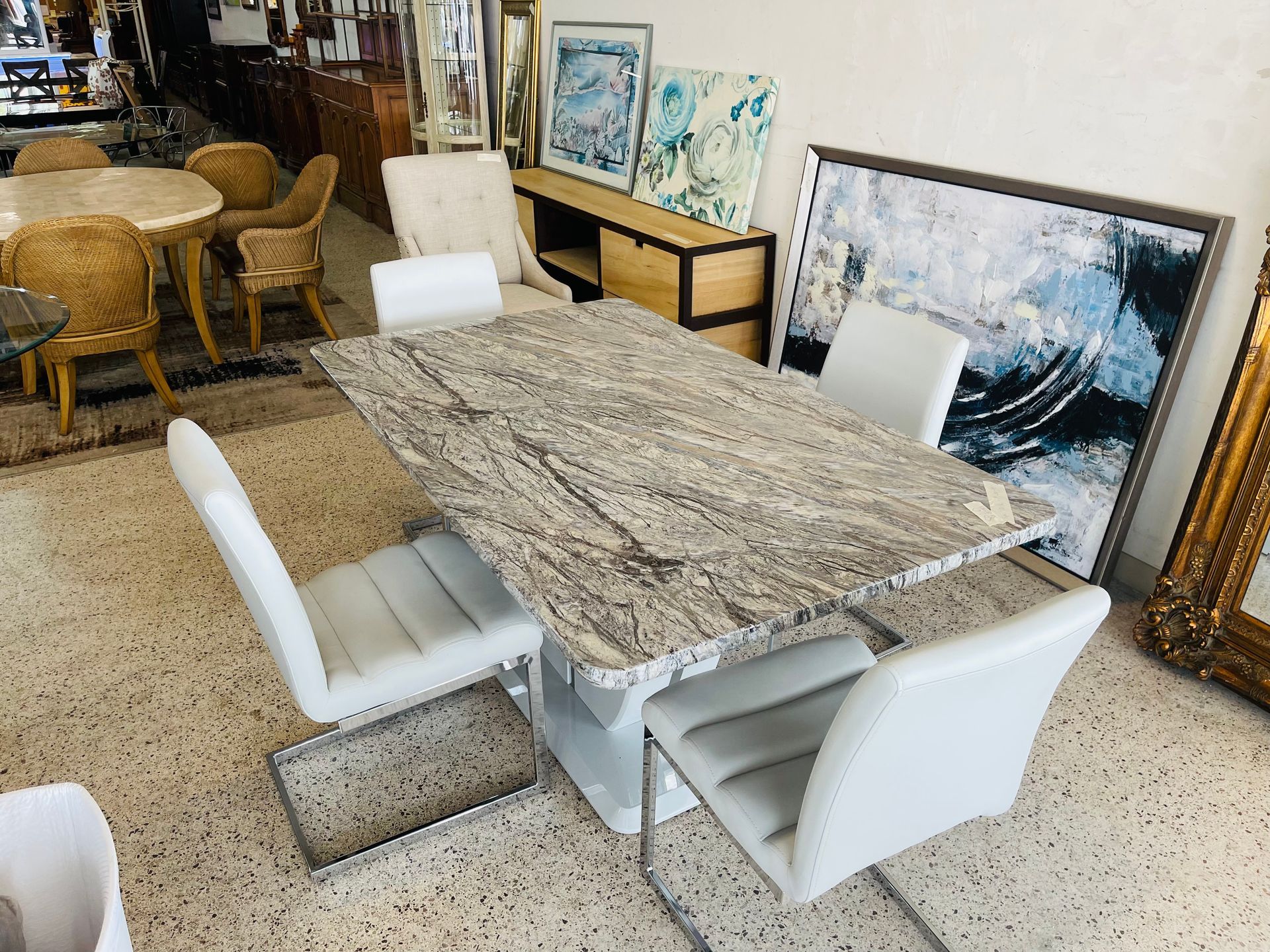 A granite dining table with four white chairs in a furniture showroom, near artwork and other chairs.