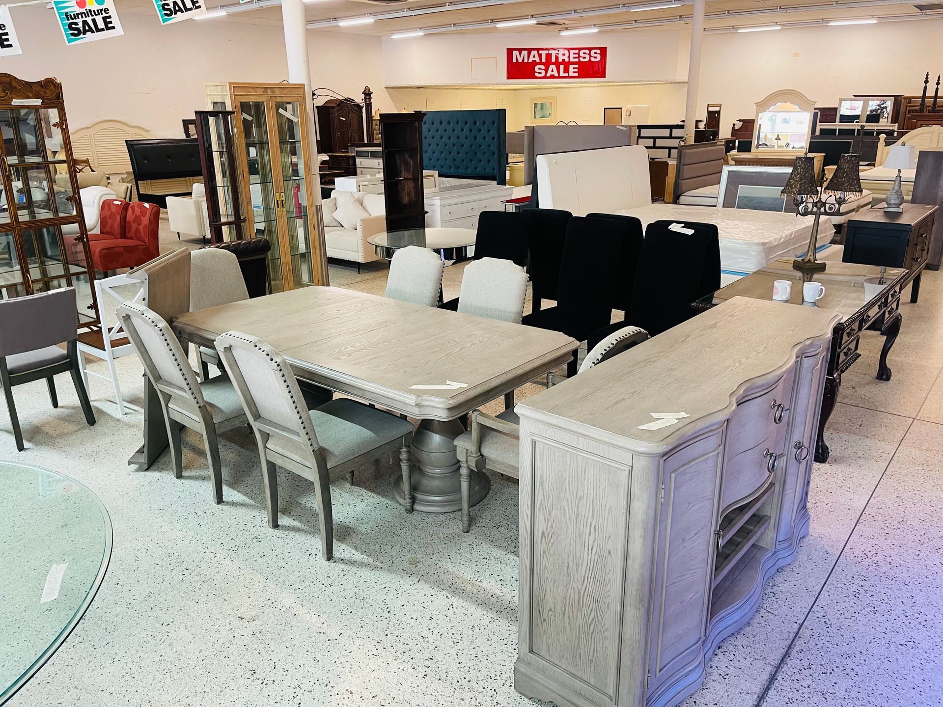 Furniture showroom floor featuring a light wood dining set, buffet, and various chairs on a speckled floor.