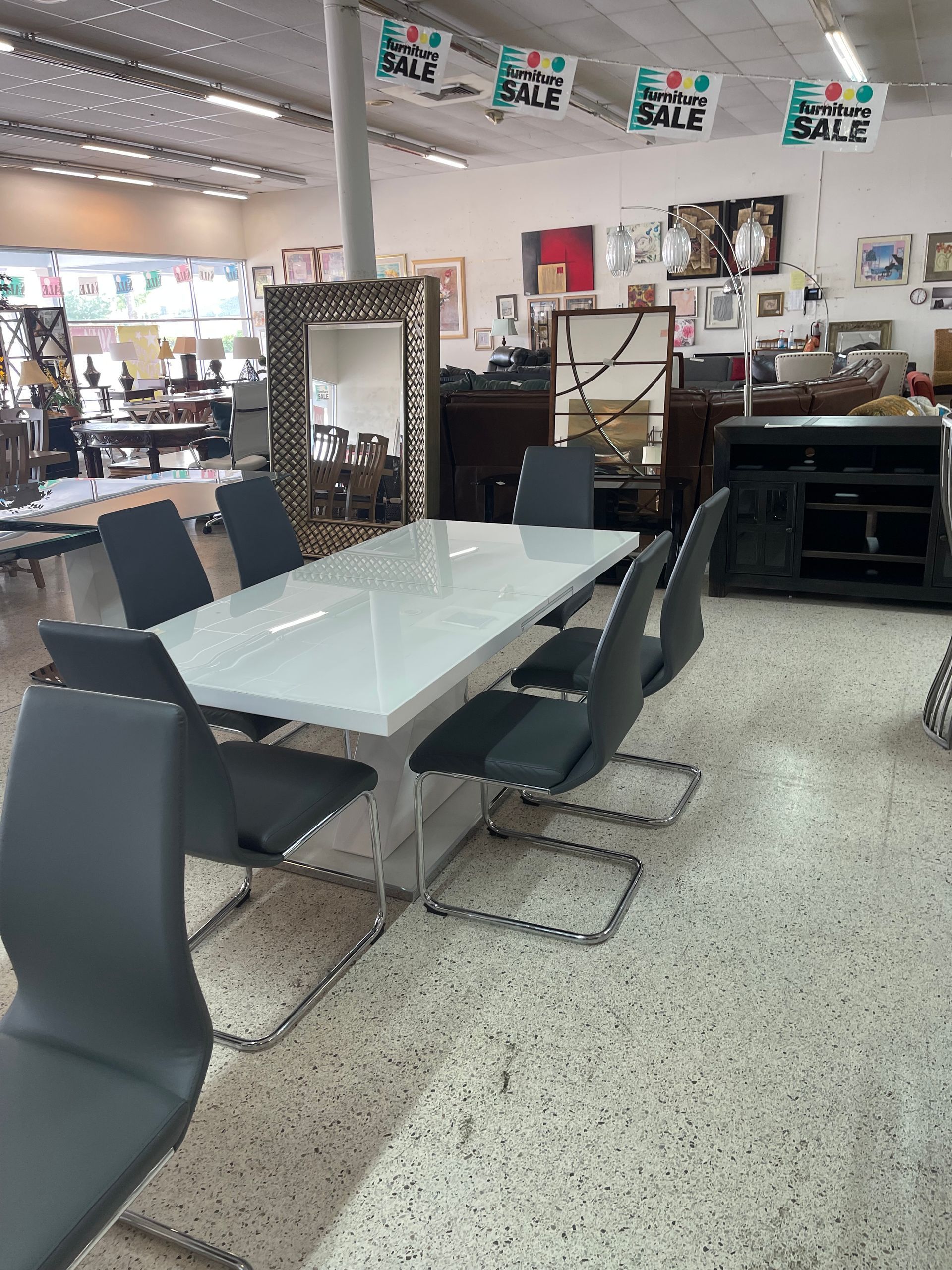 A white dining table with six gray upholstered chairs in a furniture store, with sale banners hanging from the ceiling.
